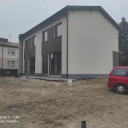 Dwukondygnacyjny dom jednorodzinny z częściowo wykończoną elewacją w kolorze beżowym i brązowym, widoczny etap prac wykończeniowych na zewnątrz, z niezagospodarowanym terenem i zaparkowanym...