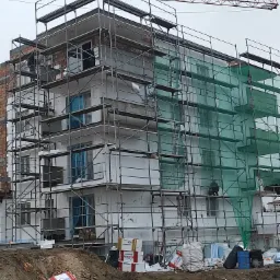 Budynek wielorodzinny w trakcie ocieplania, widoczne rusztowania, siatka ochronna i materiały izolacyjne na placu budowy, z dźwigiem w tle.