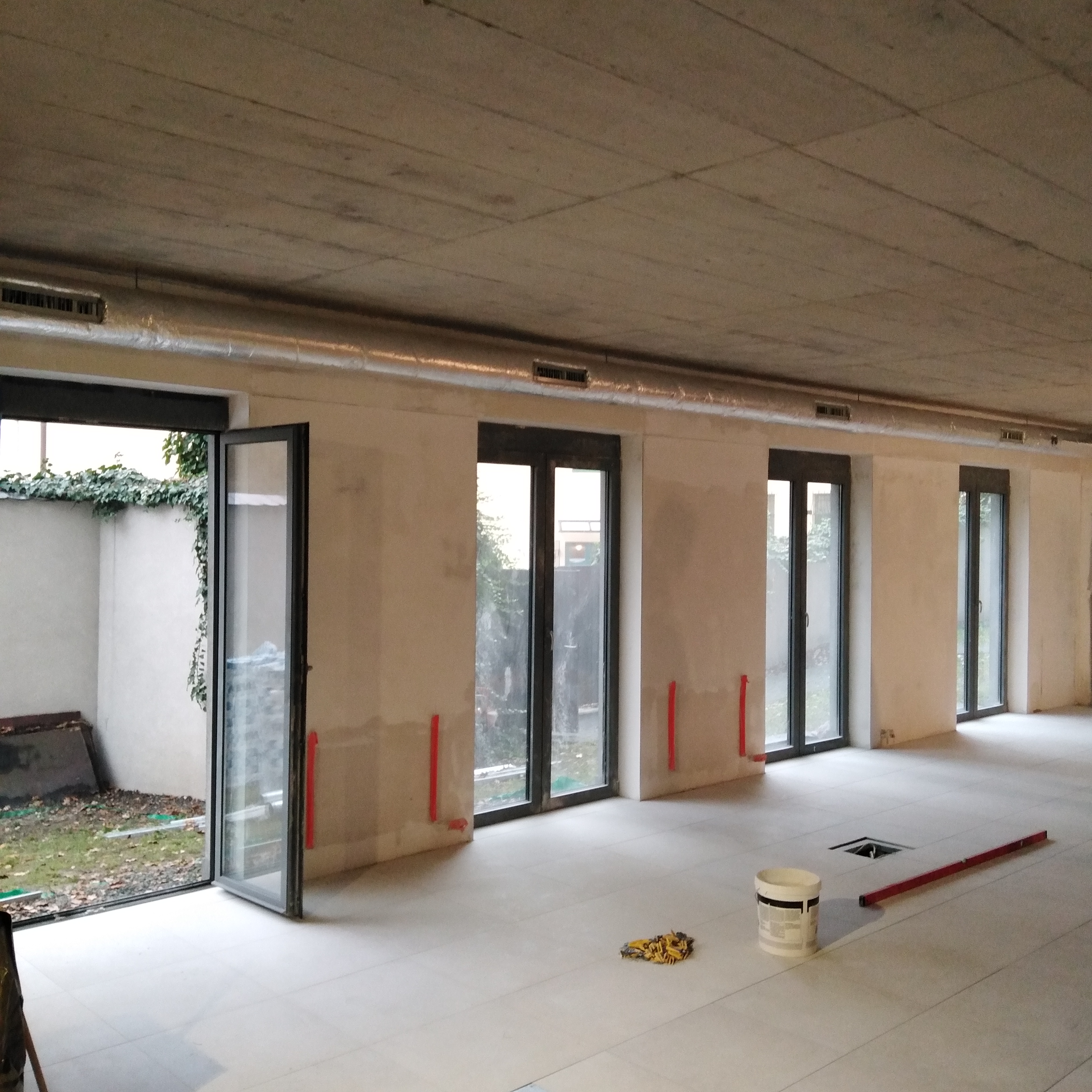 Wnętrze w trakcie prac wykończeniowych z widocznymi kanałami wentylacyjnymi pod sufitem, przygotowanymi otworami wentylacyjnymi w podłodze, oraz elementami instalacji grzewczej na ścianach.