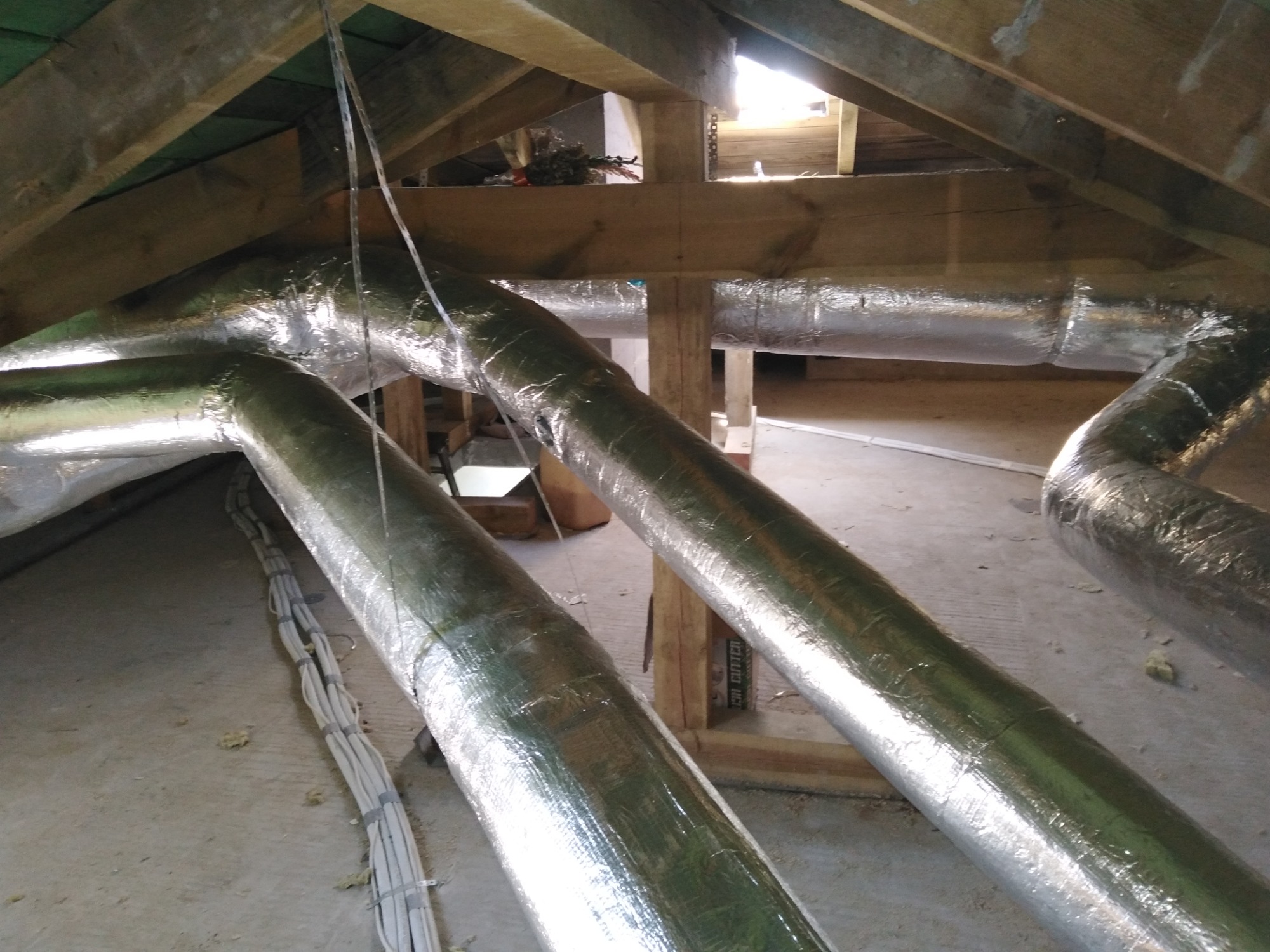 Izolowane rury wentylacyjne w przestrzeni poddasza, widoczne drewniane elementy konstrukcyjne dachu i kable elektryczne.