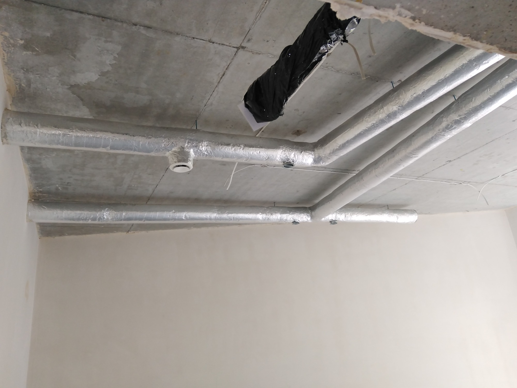 System rur wentylacyjnych z izolacją aluminiową zamontowany pod betonowym sufitem w surowym wnętrzu, widoczne elementy mocujące i czarna folia zabezpieczająca.