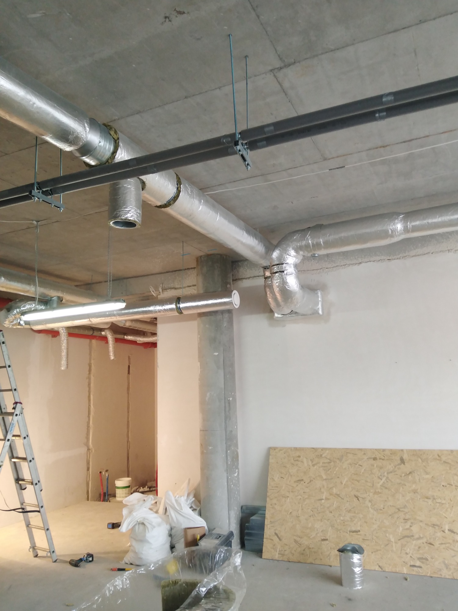 Montaż instalacji wentylacyjnej w surowym wnętrzu budynku komercyjnego, widoczne rury izolowane folią aluminiową pod betonowym sufitem, drabina i materiały budowlane na podłodze.