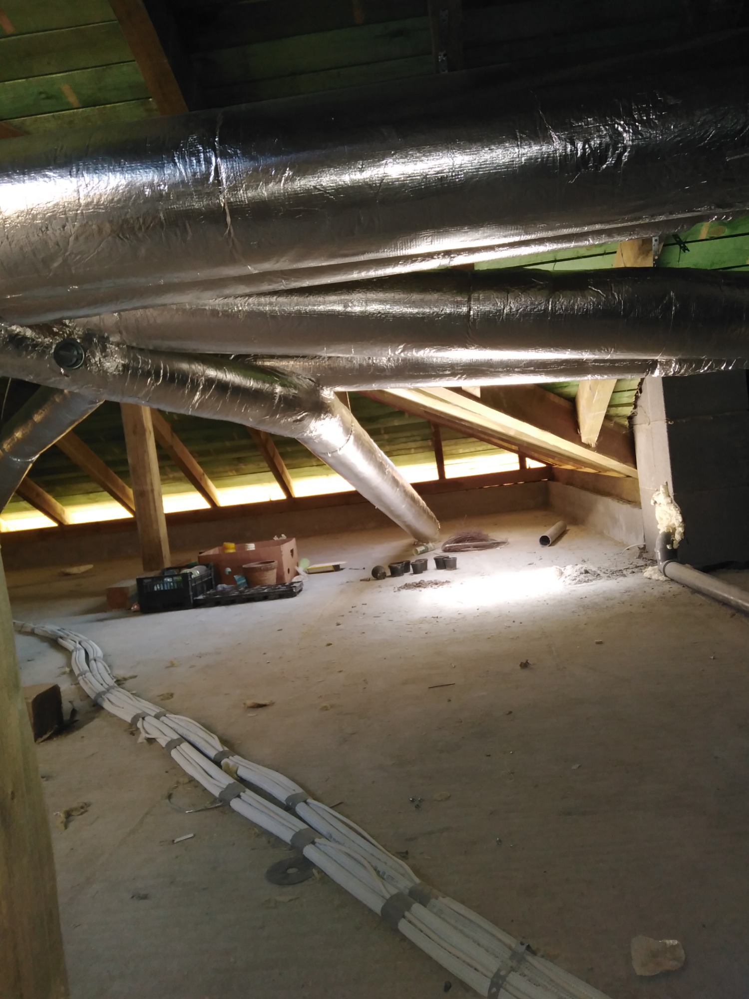 Instalacja przewodów wentylacyjnych w izolacji aluminiowej na poddaszu, widoczne elementy konstrukcji dachu i przewody elektryczne ułożone na podłodze.