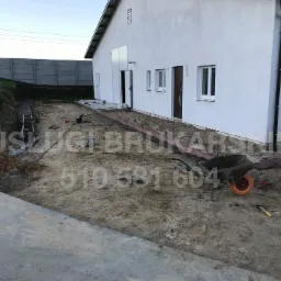 Teren budowy przy białym budynku, widoczna taczka z ziemią, przygotowane obrzeża pod kostkę, betonowy płot w tle.