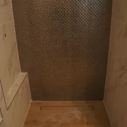 Kabiną prysznicowa z mozaiką w odcieniach brązu i beżu, z odpływem liniowym w podłodze wyłożonej płytkami imitującymi drewno. Na podłodze ślady po butach.