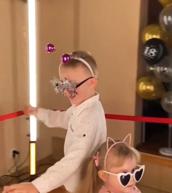 Dzieci w okularach i opaskach na głowie pozują wewnątrz, widoczne balony w tle z napisem '18'.