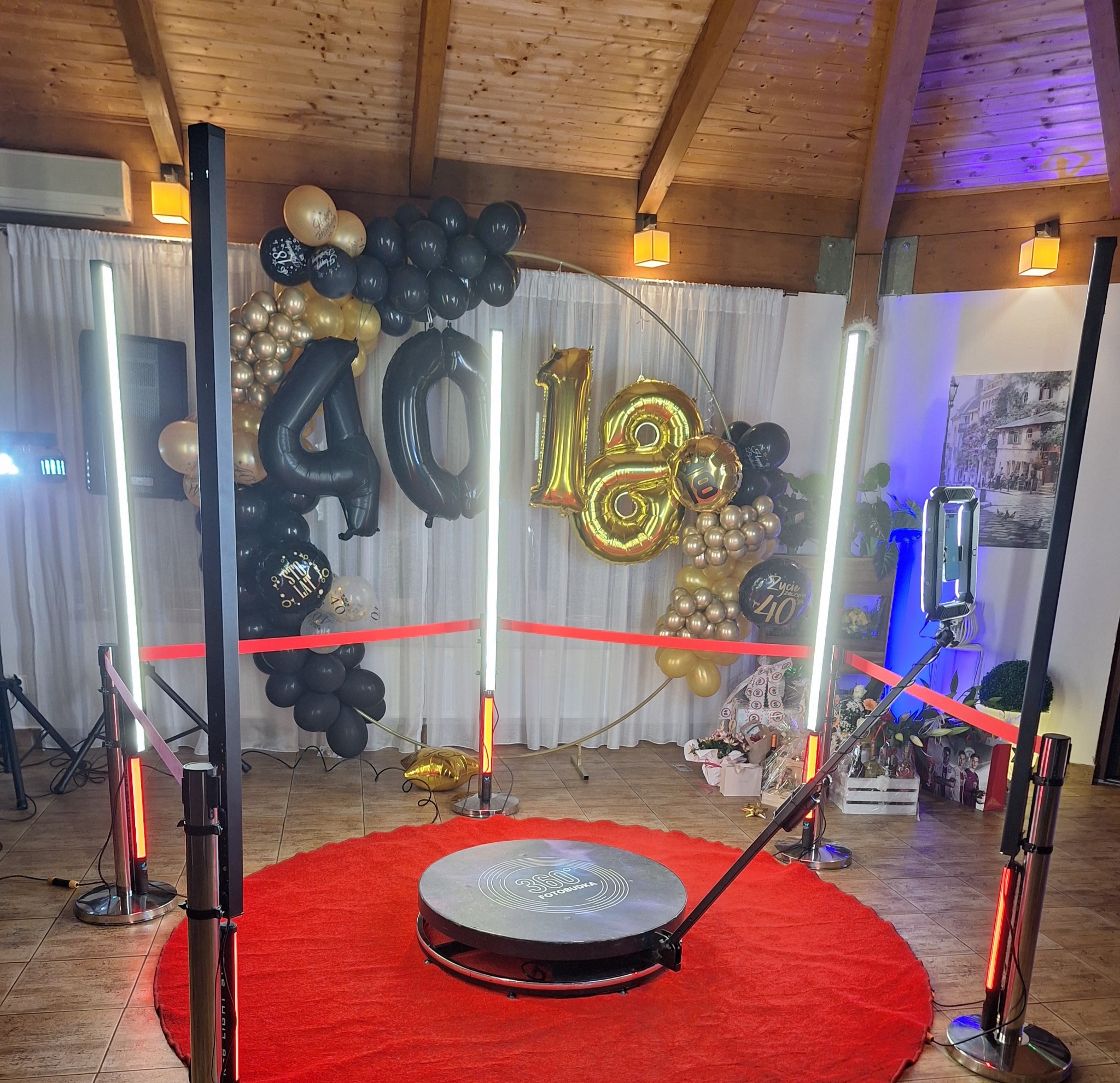 Okrągła platforma z napisem 360, otoczona czerwonym dywanem i oświetleniem LED, dekoracje balonowe w kolorach czarnym i złotym z okazji 40 i 18 urodzin w tle.