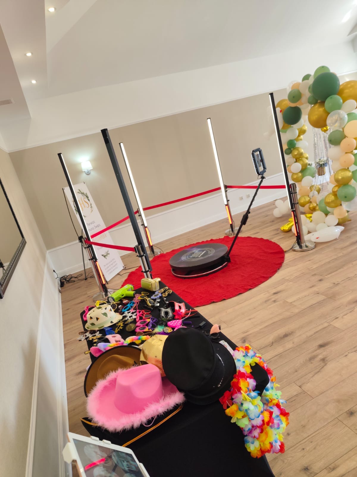 Kompletna aranżacja stanowiska fotobudki 360 z rekwizytami: kapelusze, okulary, girlandy. W tle dekoracja balonowa i czerwony dywan.