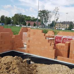 Fectumpro - Wznoszenie ścian z czerwonej cegły Porotherm na fundamencie z izolacją przeciwwilgociową, widoczne zapasy cegieł na paletach, w tle boisko sportowe i zabudowa jednorodzinna.