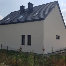 Nowoczesny dom murowany z szarą dachówką i kominami, widok z boku na elewację w jasnym odcieniu, z widocznym fragmentem ogrodzenia panelowego i sąsiedniego budynku w tle.