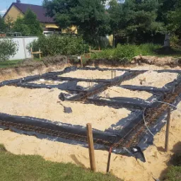 Wykonane wykopy pod fundamenty domu jednorodzinnego, zbrojenie ułożone na czarnej folii, widoczne pionowe pręty zbrojeniowe, piasek zasypowy, w tle trawa i zabudowa jednorodzinna.