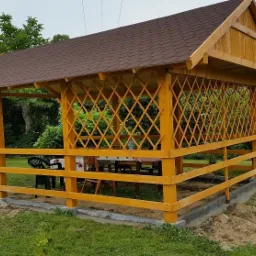 Drewniana altana z brązowym dachem z gontu bitumicznego, otoczona niskim płotkiem, z widocznymi meblami ogrodowymi wewnątrz, na tle zielonego trawnika i drzew.