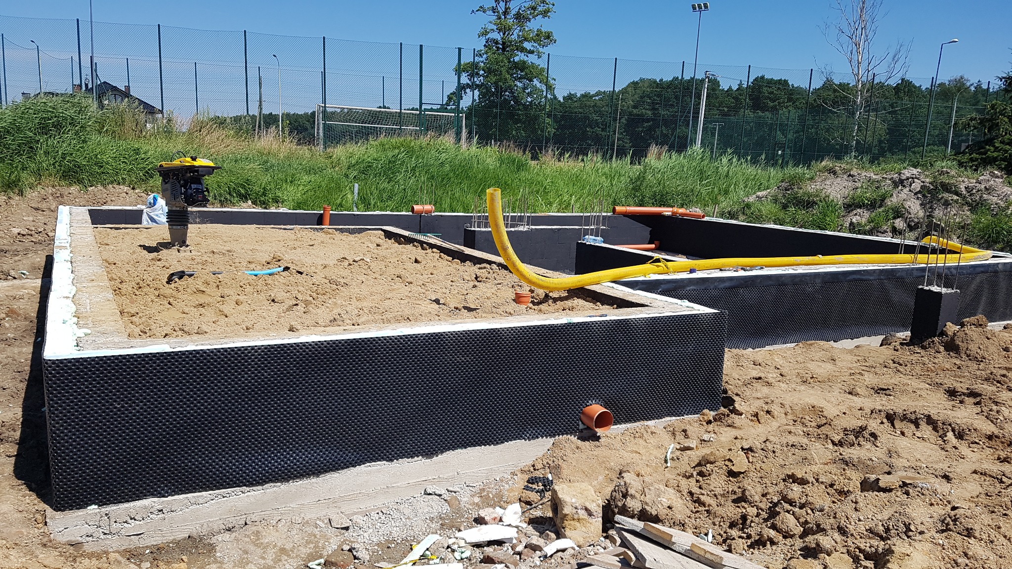 Fundamenty domu w trakcie budowy: izolacja pionowa folią kubełkową, zasypany piaskiem, żółty wąż wentylacyjny, ubijarka wibracyjna i wystające pręty zbrojeniowe na tle boiska sportowego.