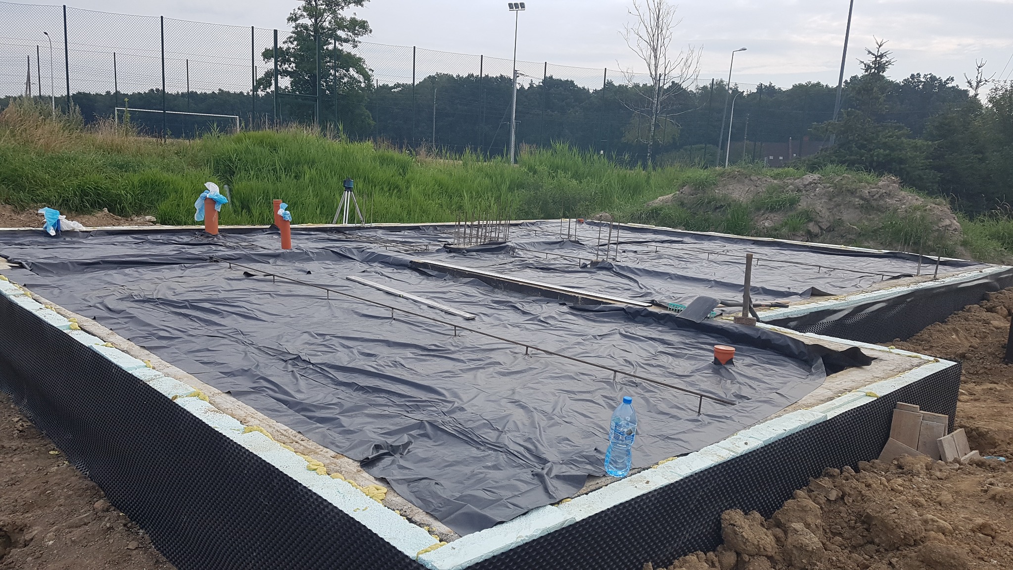Przygotowany zbrojony fundament pod budowę domu, zabezpieczony czarną folią, z wystającymi rurami instalacyjnymi i butelką wody na brzegu.