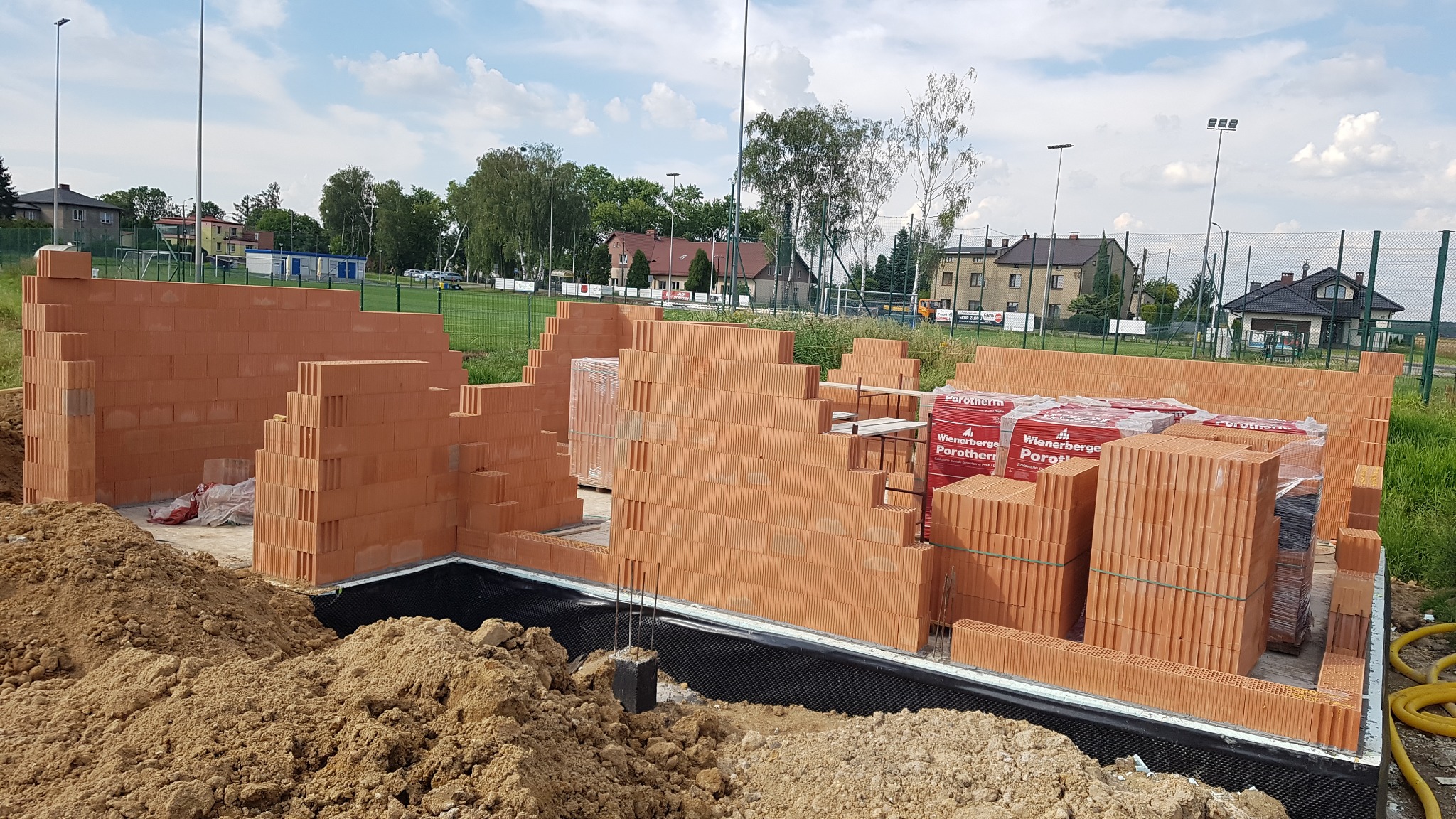 Wznoszenie ścian z czerwonej cegły Porotherm na fundamencie z izolacją przeciwwilgociową, widoczne zapasy cegieł na paletach, w tle boisko sportowe i zabudowa jednorodzinna.