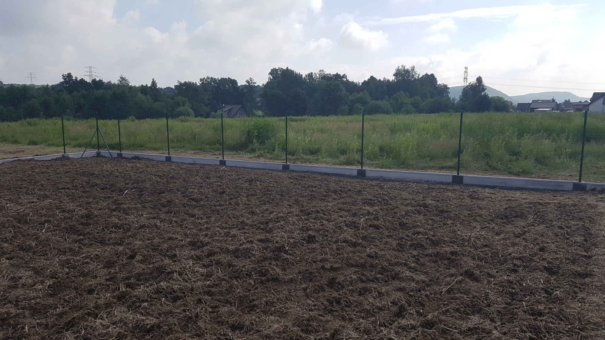 Przygotowany teren pod budowę z widocznymi fundamentami i słupkami ogrodzeniowymi, w tle zielona łąka i las.