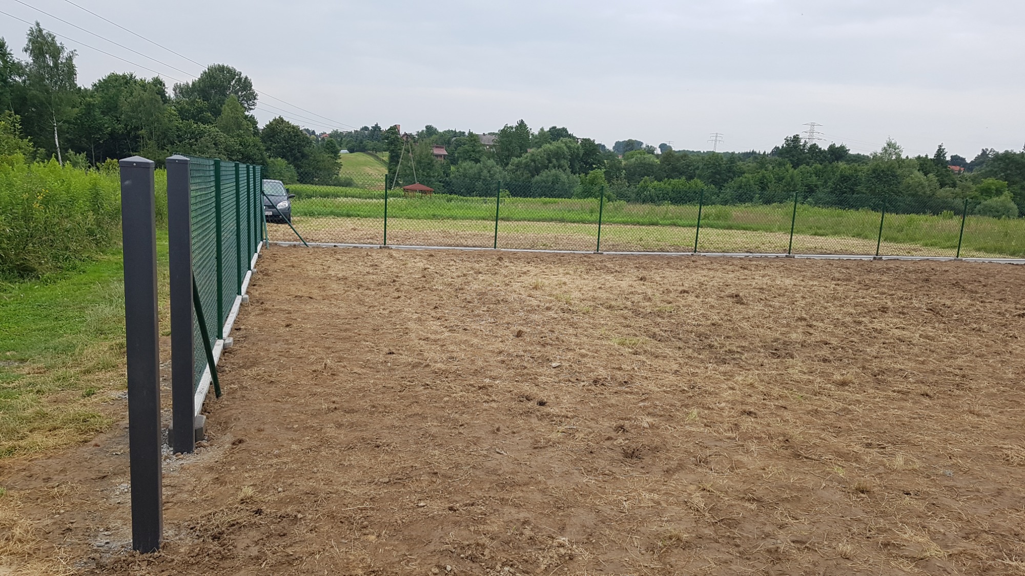Nowo postawione ogrodzenie z siatki w kolorze zielonym z czarnymi słupkami na przygotowanym terenie, w tle widoczny krajobraz z drzewami i domami.