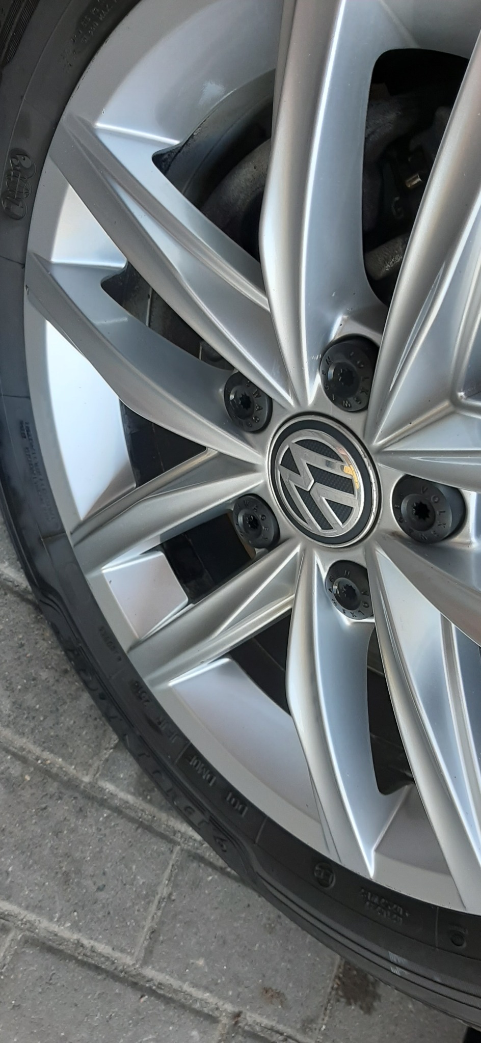 Srebrna felga aluminiowa z oponą na tle kostki brukowej, widoczne logo Volkswagen na deklu śruby.