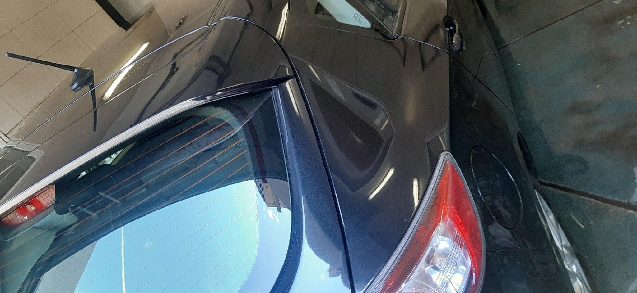 Czarny samochód po umyciu, widoczny fragment tylnej klapy z lampą i szybą, na lakierze widoczne odbicia świetlówek.