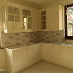 Jasna, narożna zabudowa kuchenna z białymi frontami, szklanymi witrynami w górnych szafkach i płytkami w kratkę nad blatem. Brak widocznego sprzętu AGD.