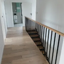 Nowoczesne schody z drewnianymi stopniami i czarną, metalową balustradą w jasnym wnętrzu z drewnianą podłogą. W tle widoczna łazienka w trakcie wykańczania.