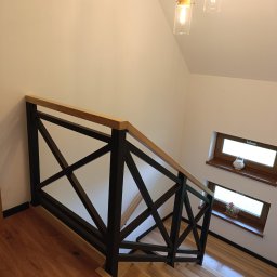 AKARDOM - Nowoczesna balustrada schodowa z czarnymi, krzyżującymi się elementami i jasnym drewnianym poręczą, oświetlona przez wiszące lampy. Widok z góry na drewniane schody.