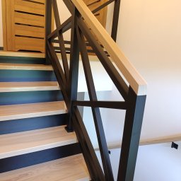 AKARDOM - Nowoczesne schody z jasnego drewna z czarnymi podstopnicami i geometryczną, metalową balustradą z drewnianą poręczą, widok z dołu, drzwi w tle.