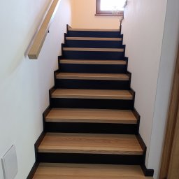 AKARDOM - Nowoczesne schody drewniane z czarnymi podstopnicami i jasną poręczą, widok od dołu. Minimalistyczny design, naturalne światło z okna na poddaszu.