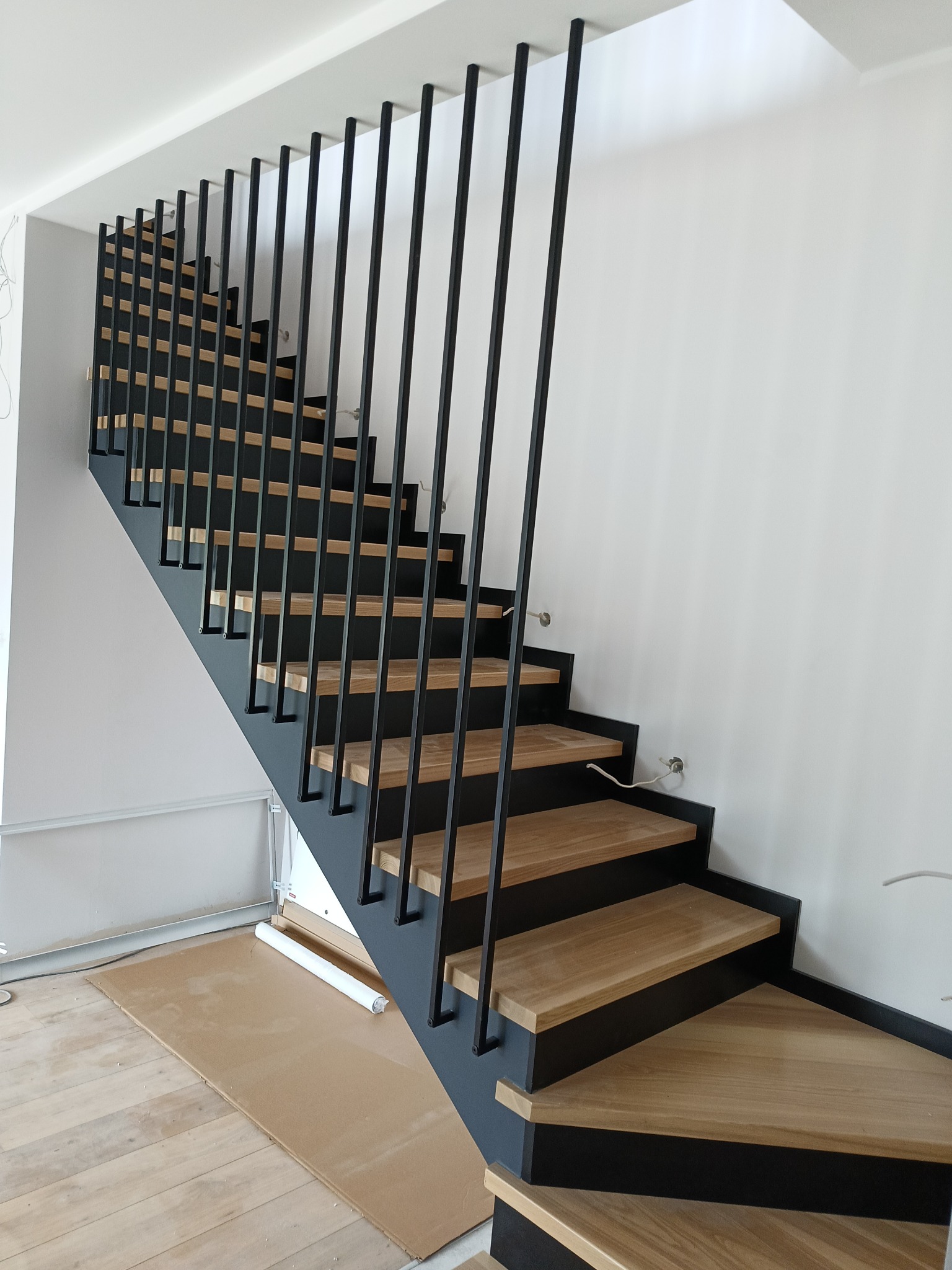 Nowoczesne schody z drewnianymi stopniami i czarną metalową balustradą ze szczebelkami, widoczne przewody elektryczne wystające ze ściany, zabezpieczenie podłogi kartonem.