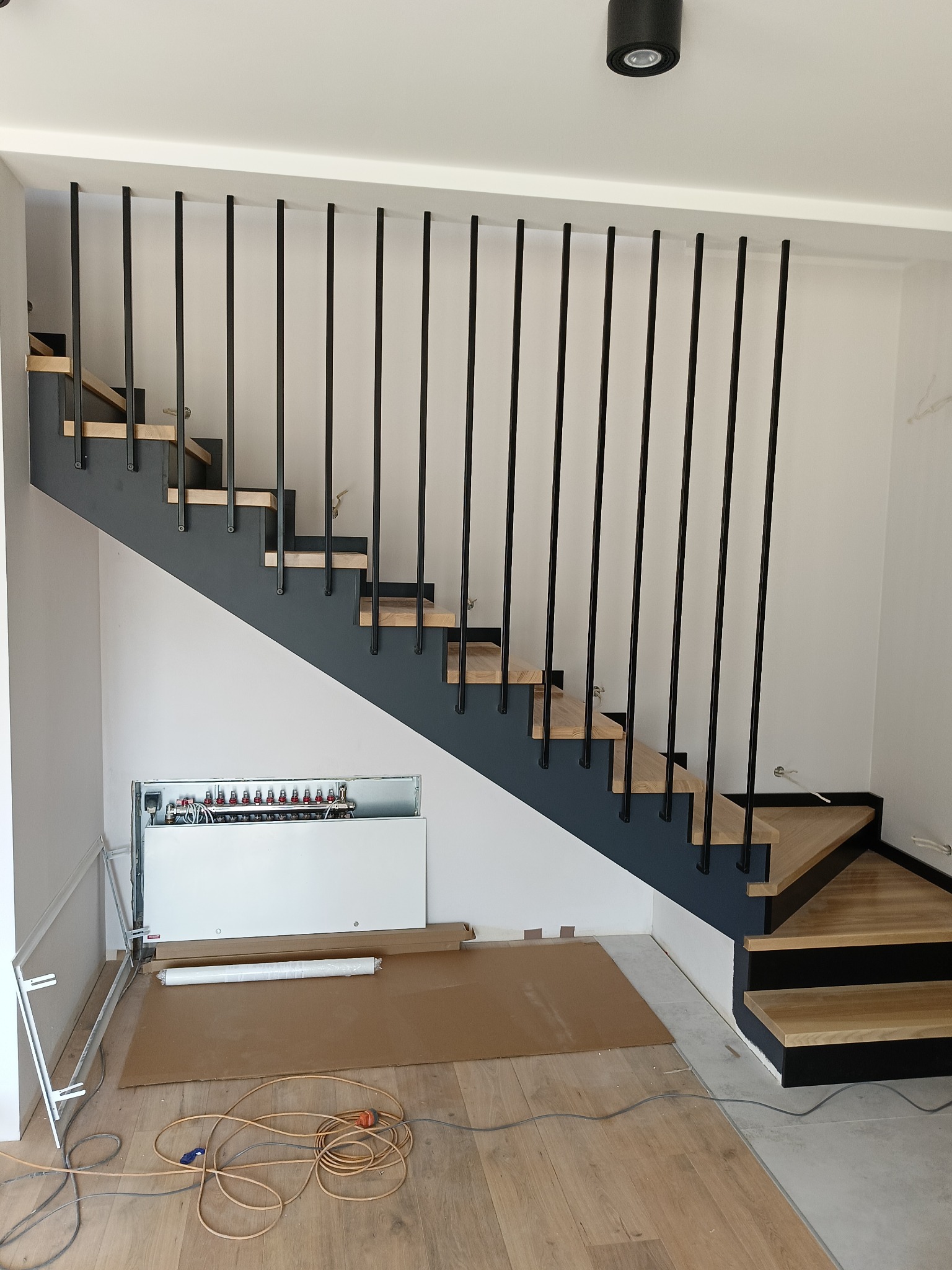 Nowoczesne schody z drewnianymi stopniami, czarną konstrukcją i minimalistycznymi balustradami z pionowych, czarnych prętów. Widoczna instalacja grzewcza pod schodami i materiały budowlane...