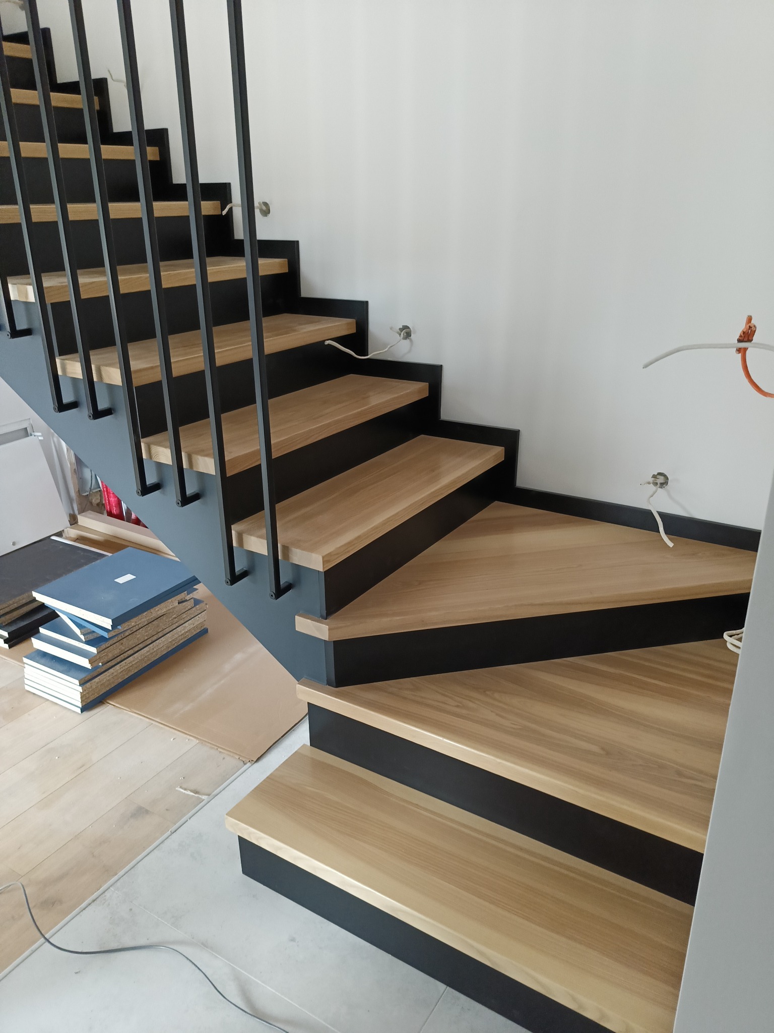 Nowoczesne, drewniane schody z czarną metalową balustradą, widoczne niedokończone prace wykończeniowe, w tle ułożone panele podłogowe i wystające kable elektryczne.