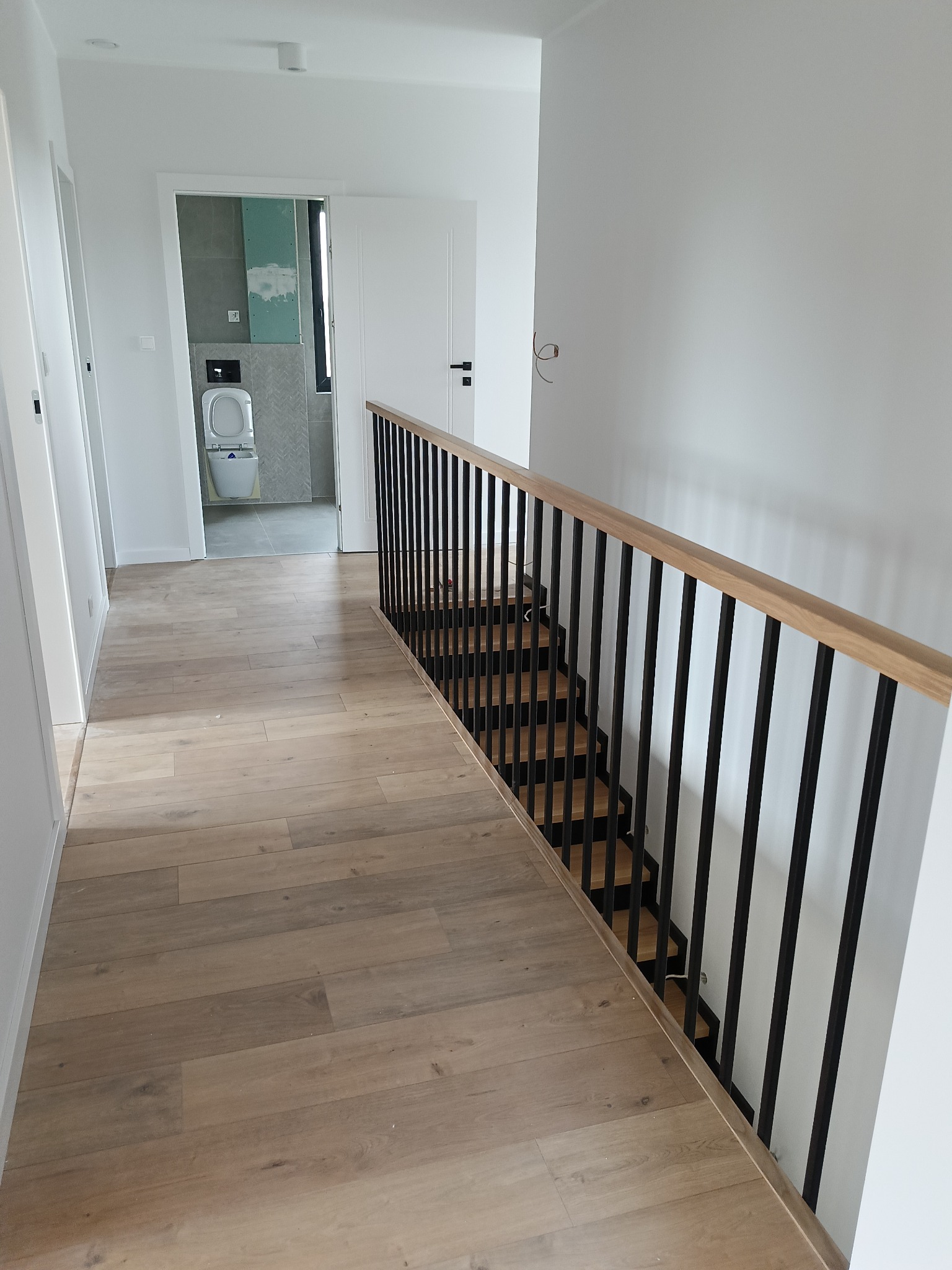 Nowoczesne schody z drewnianymi stopniami i czarną, metalową balustradą w jasnym wnętrzu z drewnianą podłogą. W tle widoczna łazienka w trakcie wykańczania.