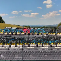 Rząd szarych modułów Grenton Relay z podłączonymi niebieskimi i czarnymi kablami na tle zielonego pola i gór z błękitnym niebem