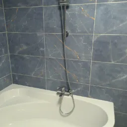 Biała wanna narożna z baterią i słuchawką prysznicową na tle ściany wyłożonej ciemnymi, marmurkowymi płytkami z delikatnymi złotymi żyłkami.