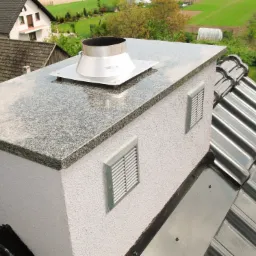 Wykończony komin z szarym, nakrapianym blatem, metalowym wkładem i kratkami wentylacyjnymi na tle dachu i zieleni.