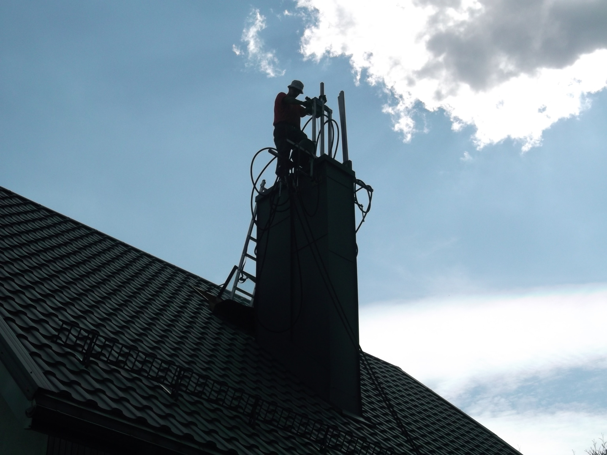 Pracownik w kasku na dachu, instalujący antenę na kominie z wykorzystaniem drabiny, na tle błękitnego nieba z chmurami.