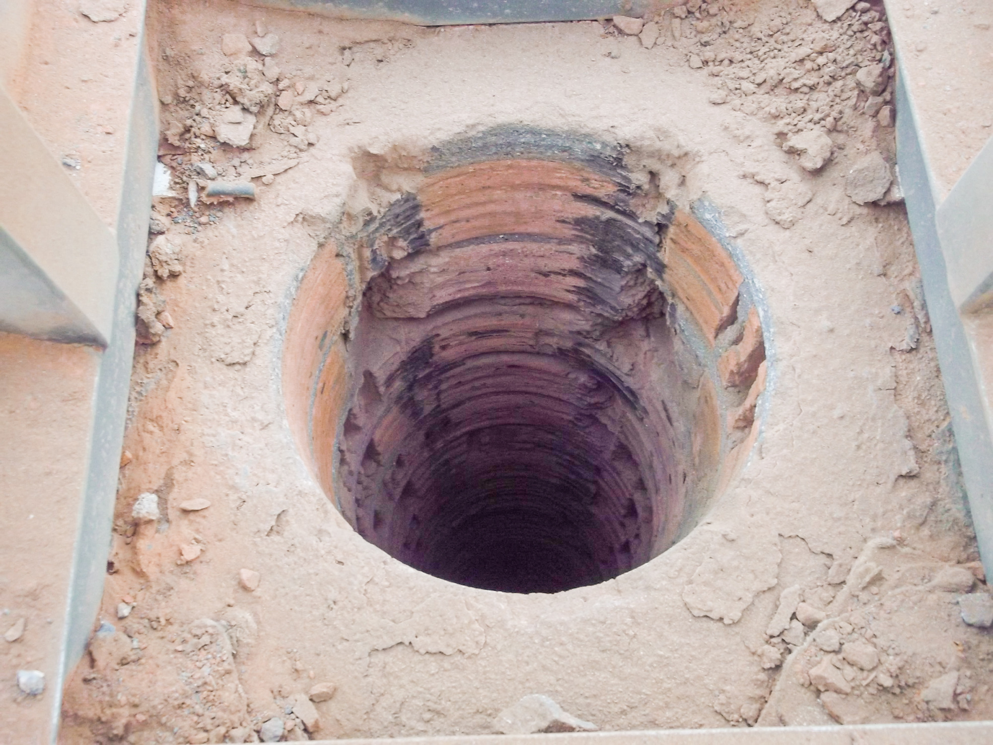 Otwór w ziemi wyłożony cegłą, prawdopodobnie fragment komina w trakcie budowy lub naprawy, otoczony ziemią i elementami konstrukcyjnymi.