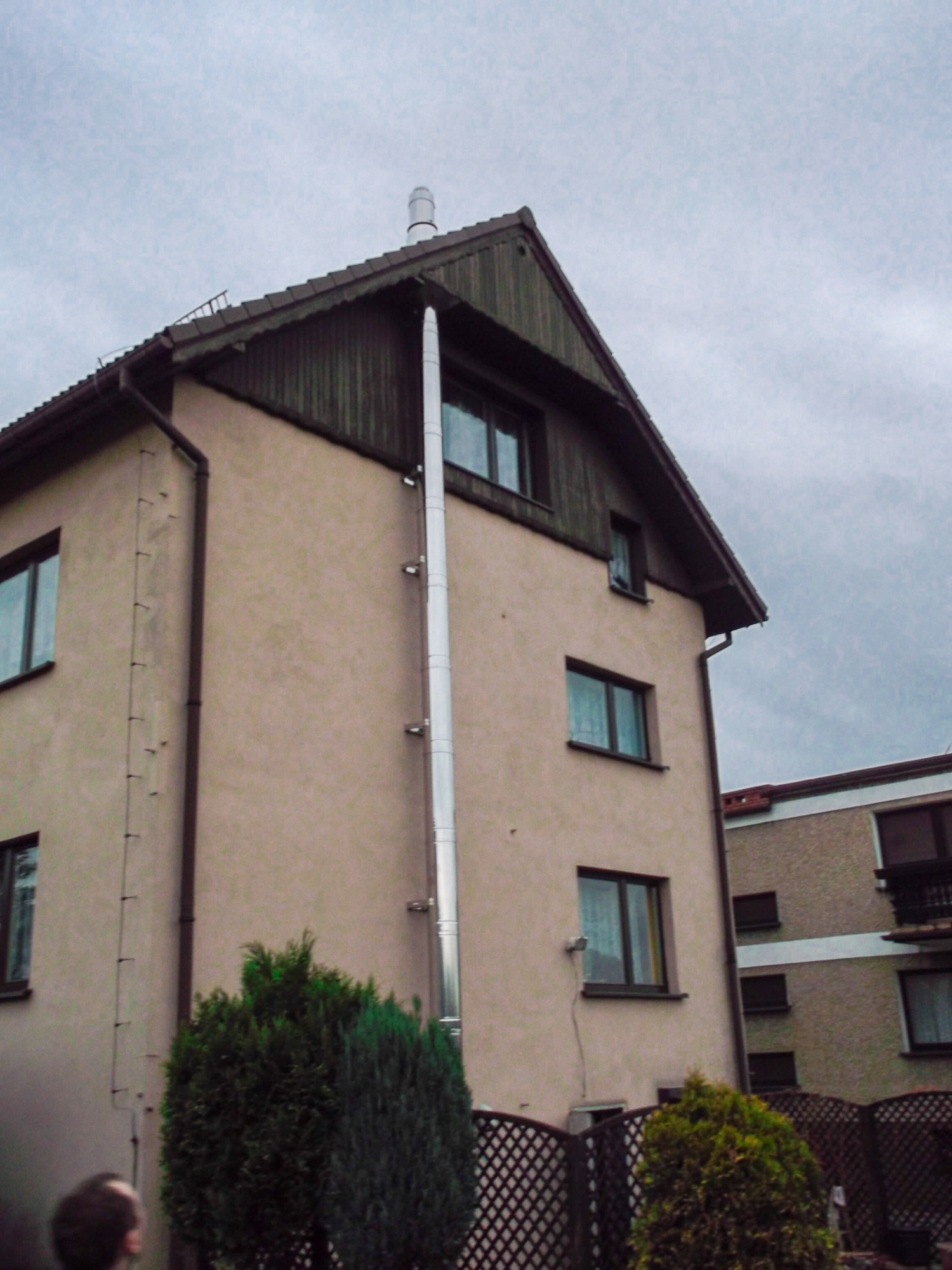 Zewnętrzny, metalowy komin zamontowany na elewacji dwupiętrowego budynku mieszkalnego, widoczny z poziomu gruntu.