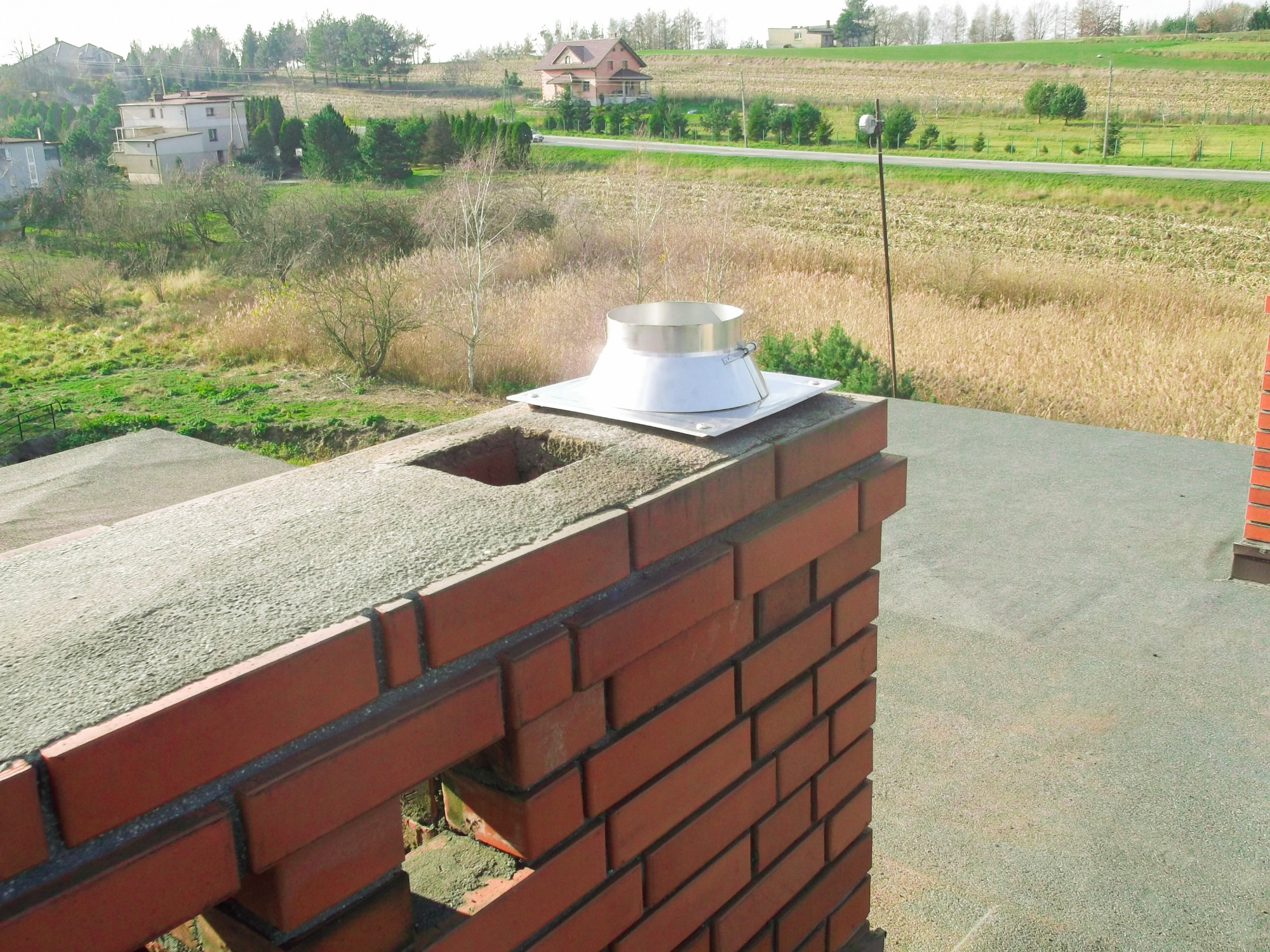 Murowany komin z zamontowanym, metalowym wkładem kominowym na tle krajobrazu wiejskiego z domami i polami.