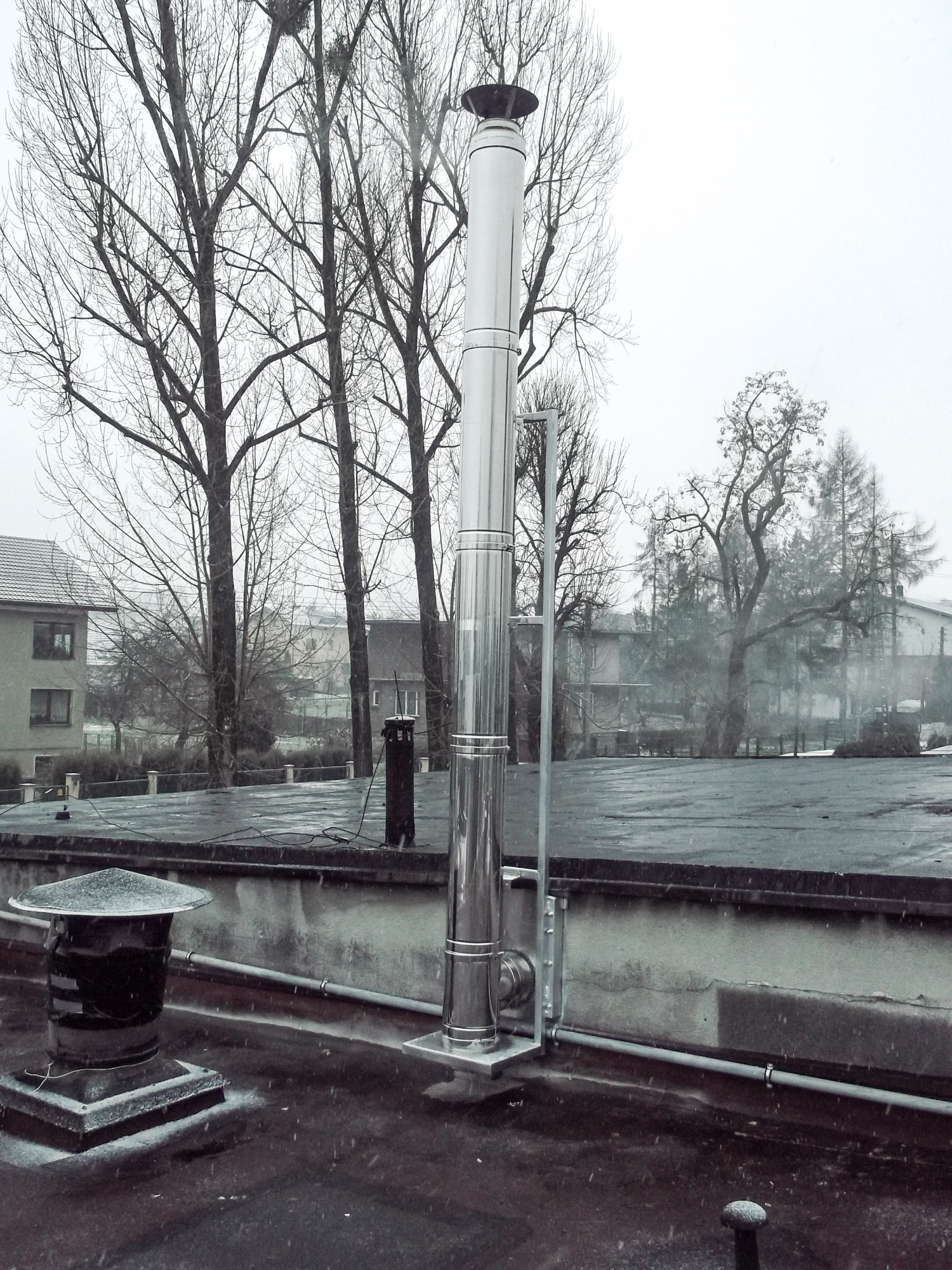 Montaż nowego, błyszczącego, stalowego przewodu kominowego na dachu pokrytym papą, w tle drzewa bez liści i budynki mieszkalne podczas opadów śniegu.