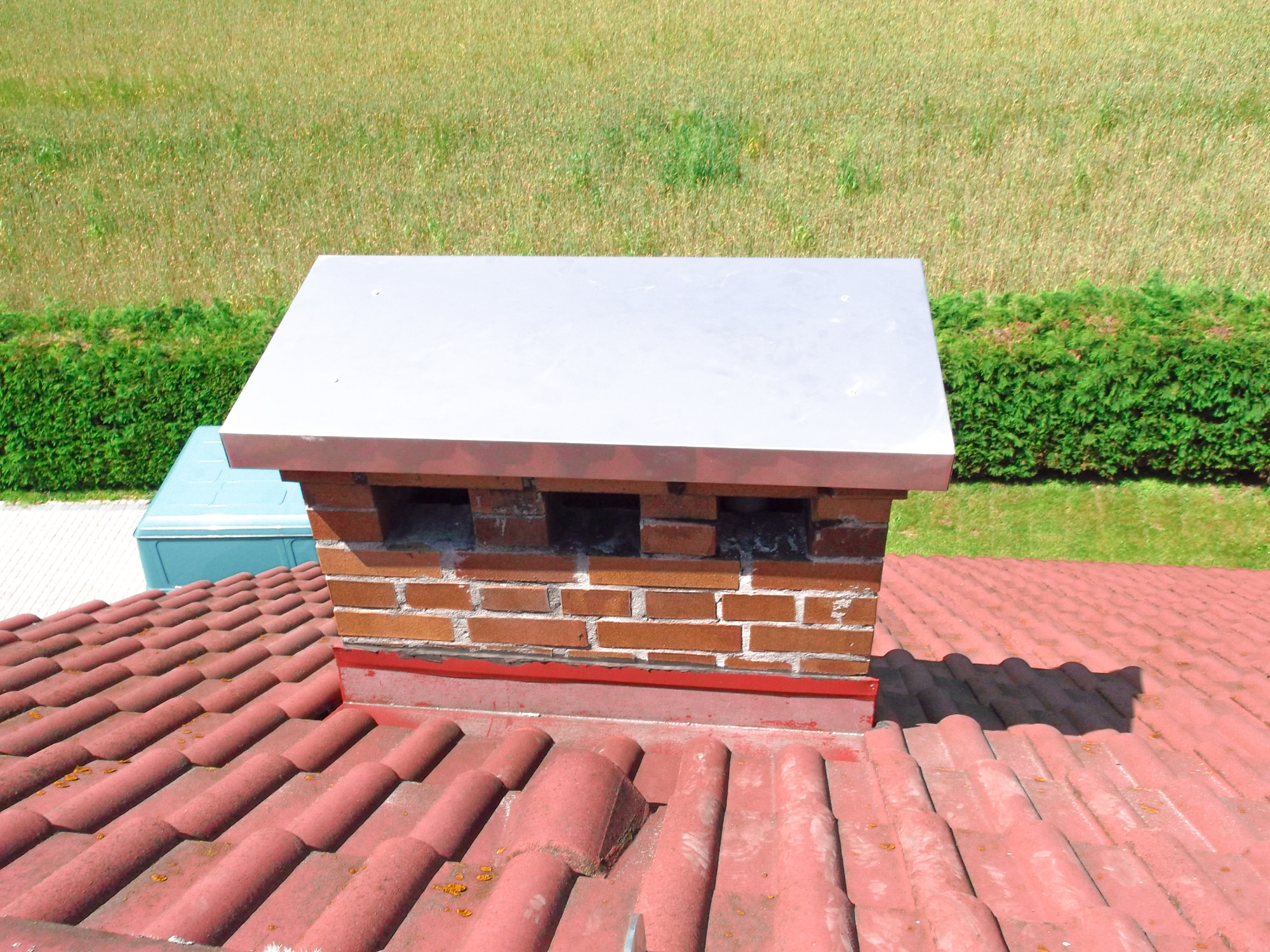 Ceglany komin z metalowym daszkiem na czerwonym dachu, widok z góry na tle zieleni.