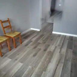 Ułożone panele podłogowe w kolorze szaro-beżowym, widoczne dwa drewniane krzesła z poduszkami w paski, szare ściany i białe listwy przypodłogowe.