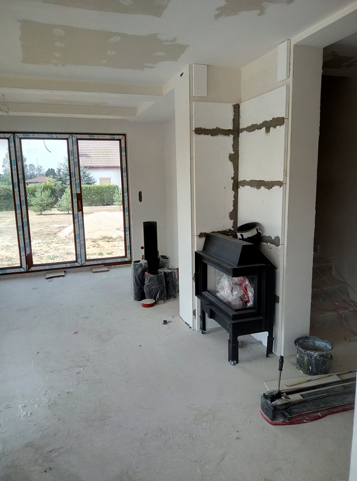 Wnętrze w trakcie wykończenia z kominkiem w obudowie z płyt gipsowych, widoczne zaszpachlowane łączenia płyt, okno z brązowymi ramami i widokiem na ogród.