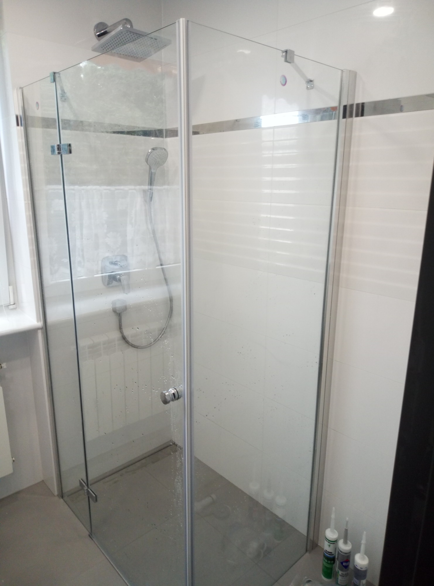 Kabina prysznicowa z przezroczystego szkła, z chromowanymi okuciami, zamontowana w łazience wyłożonej białymi płytkami; widoczne ślady wody na szkle i tuby z silikonem uszczelniającym przy ścianie.