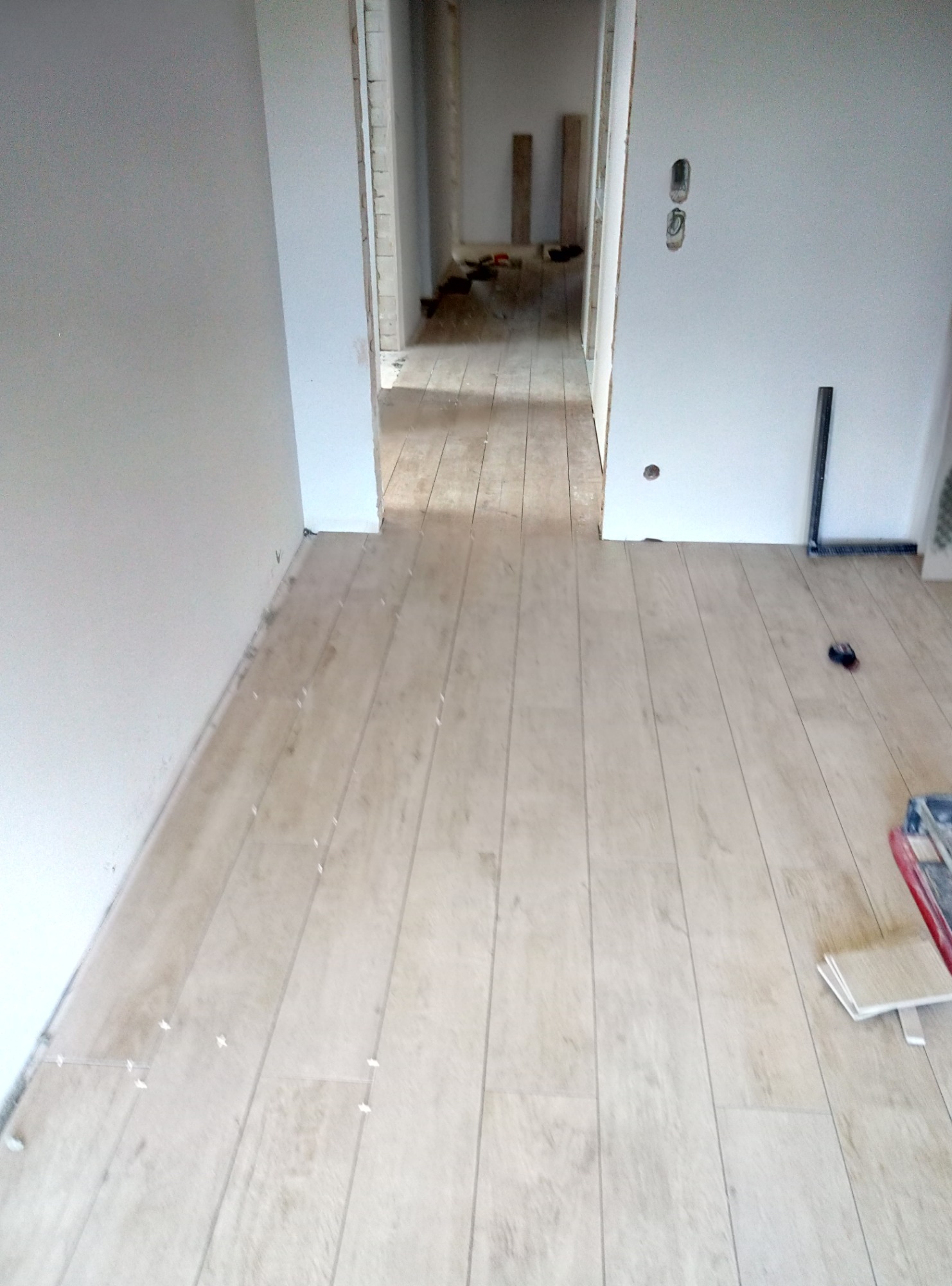 Układanie paneli podłogowych w jasnym odcieniu drewna, widoczne krzyżyki dystansowe, narzędzia i pozostałości po pracy w tle.