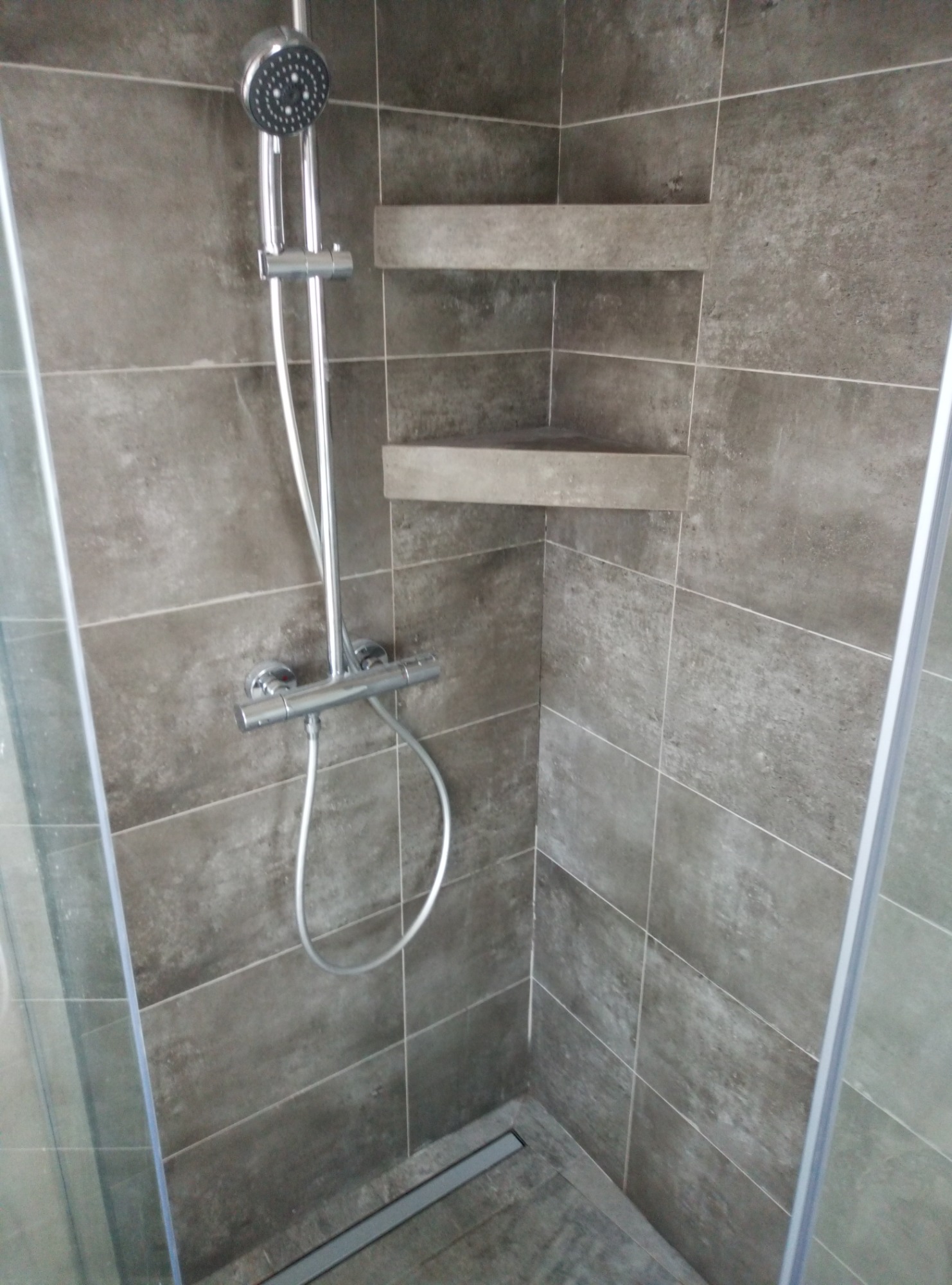 Szczegółowe ujęcie kabiny prysznicowej z szarymi płytkami, wbudowanymi półkami i nowoczesną baterią, widok z boku przez szklaną ściankę.