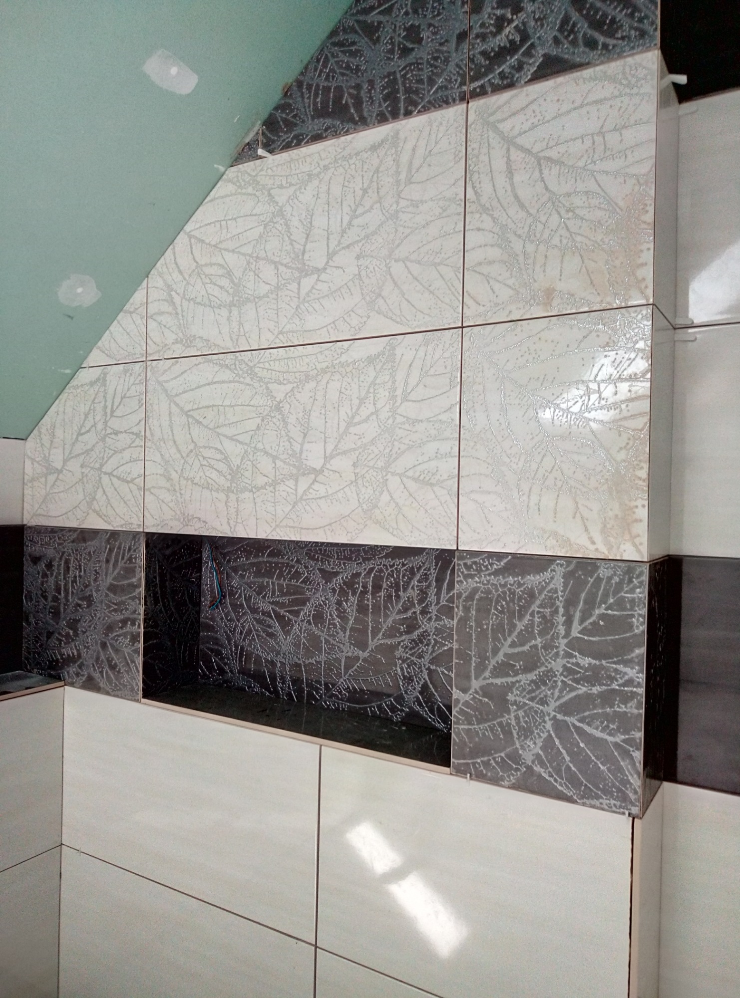 Ściana łazienki wyłożona błyszczącymi, kwadratowymi płytkami w dwóch odcieniach z motywem liści, z wnęką w dolnej części, widoczne niedokończone wykończenie sufitu z płyt gipsowo-kartonowych.