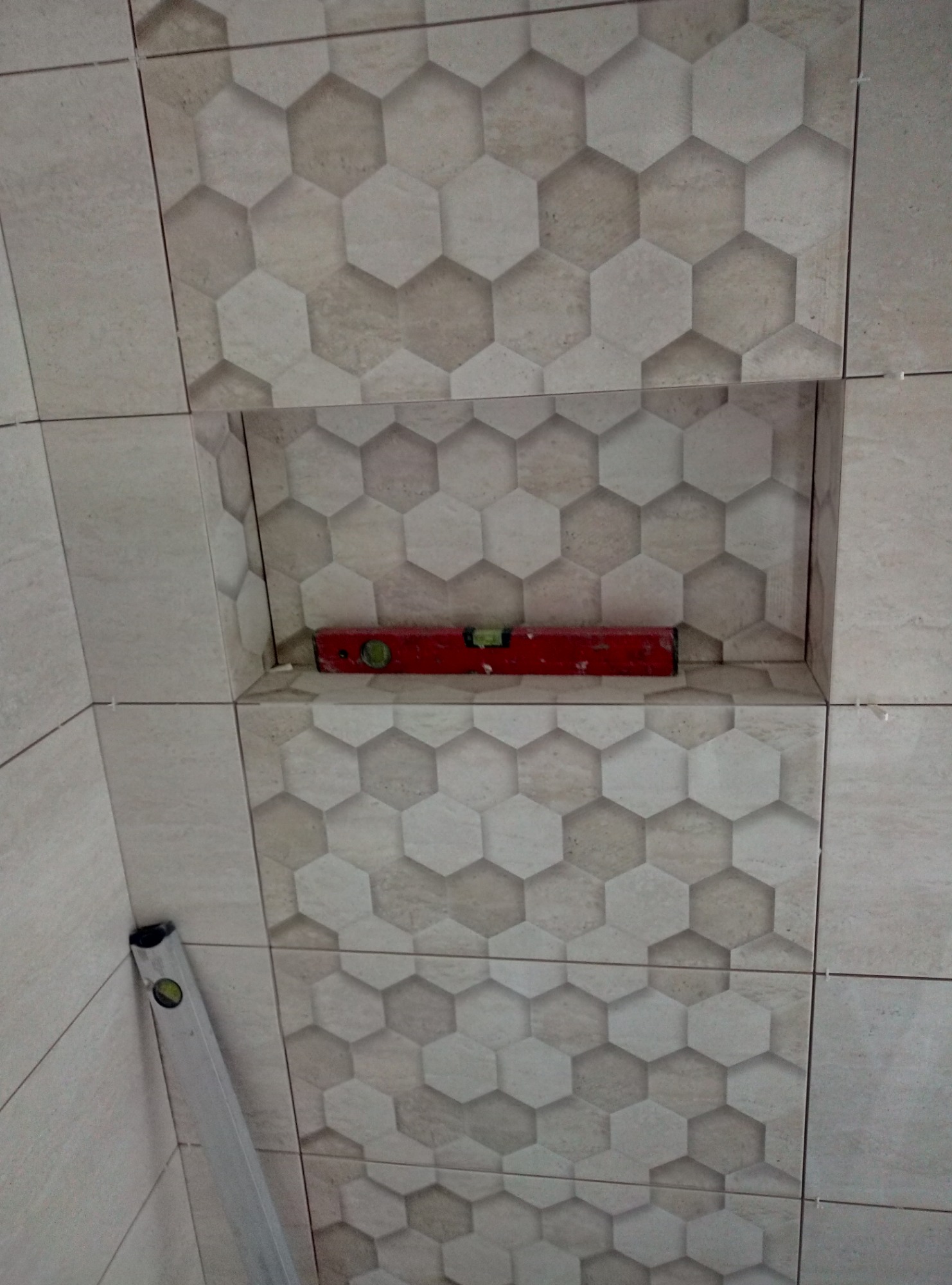 Ściana łazienki wyłożona płytkami heksagonalnymi w odcieniach beżu i szarości, z wnęką, w której umieszczono poziomicę w kolorze czerwonym, a obok ściany stoi długa poziomica aluminiowa.