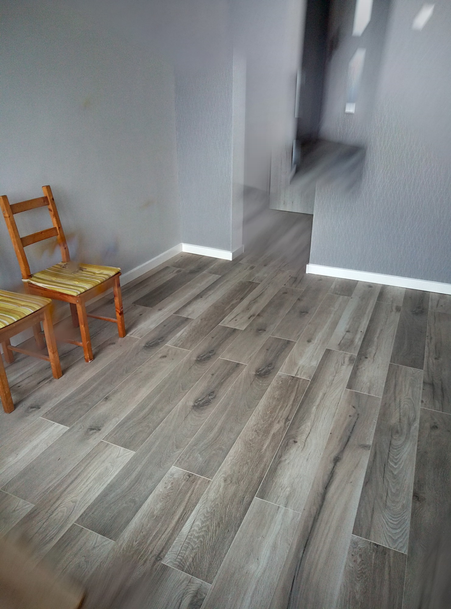 Ułożone panele podłogowe w kolorze szaro-beżowym, widoczne dwa drewniane krzesła z poduszkami w paski, szare ściany i białe listwy przypodłogowe.