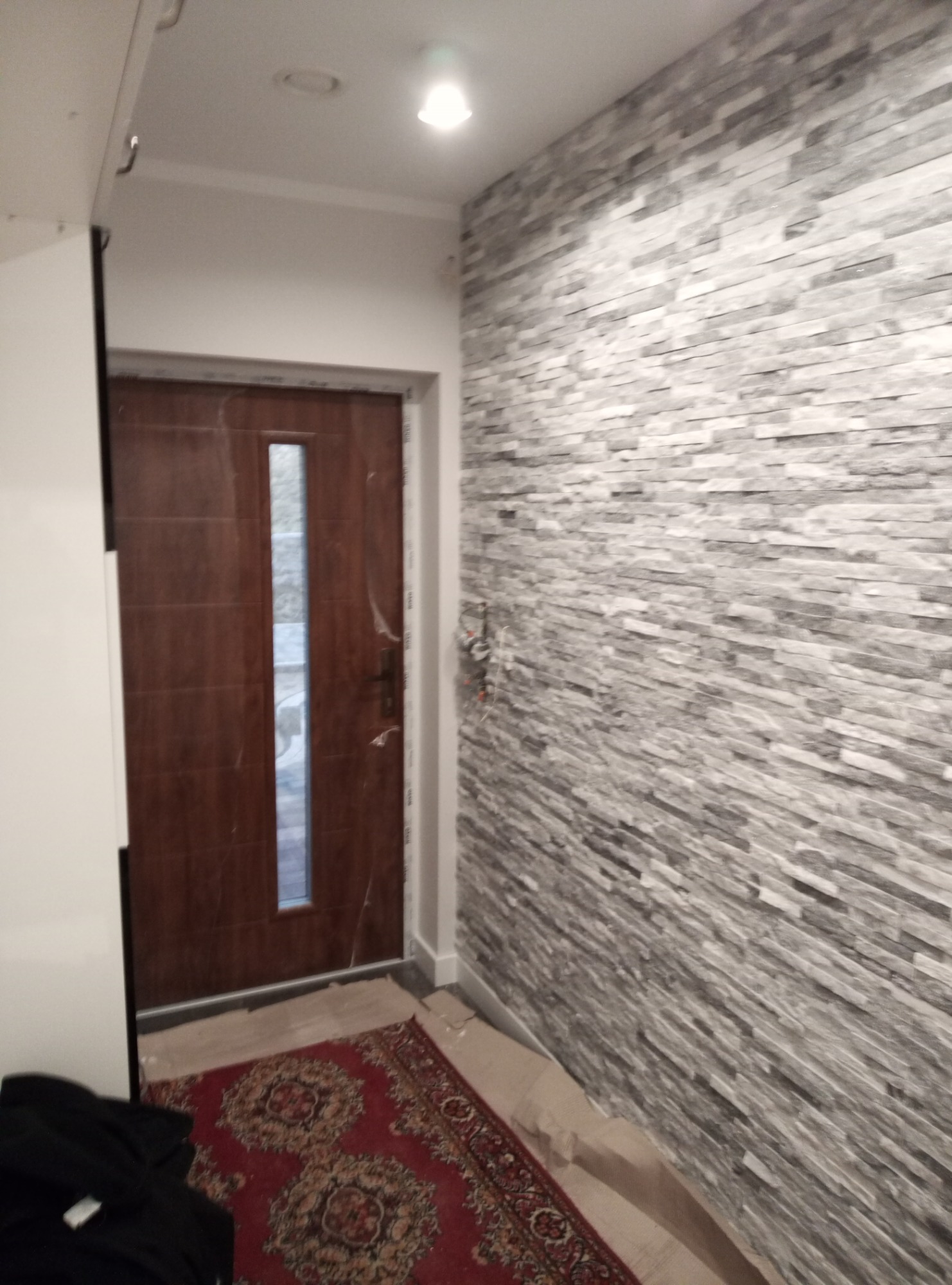 Wnętrze przedpokoju z dekoracyjną ścianą z szarej cegły, brązowymi drzwiami z szybą, dywanem w orientalny wzór i widocznymi elementami instalacji elektrycznej.